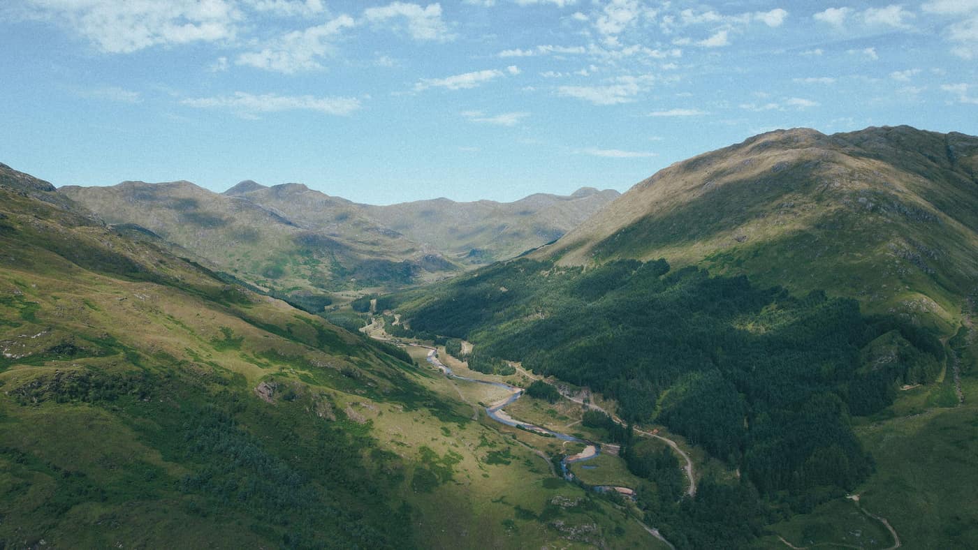 A wide Highland glen seen from above, forested slopes falling to a river, with rounded summits ranged beyond.