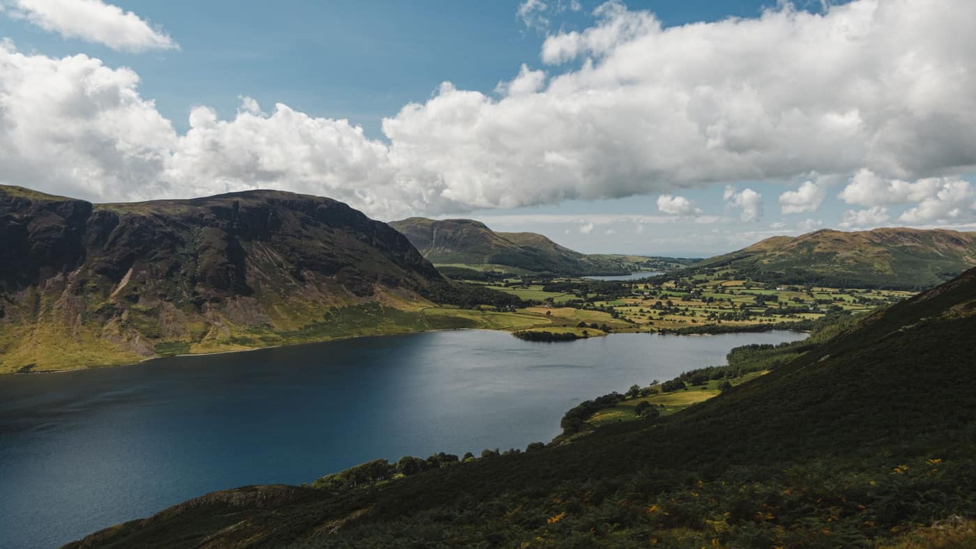 A long lake below green Lakeland fells, with hedged fields and scattered trees on the far shore.
