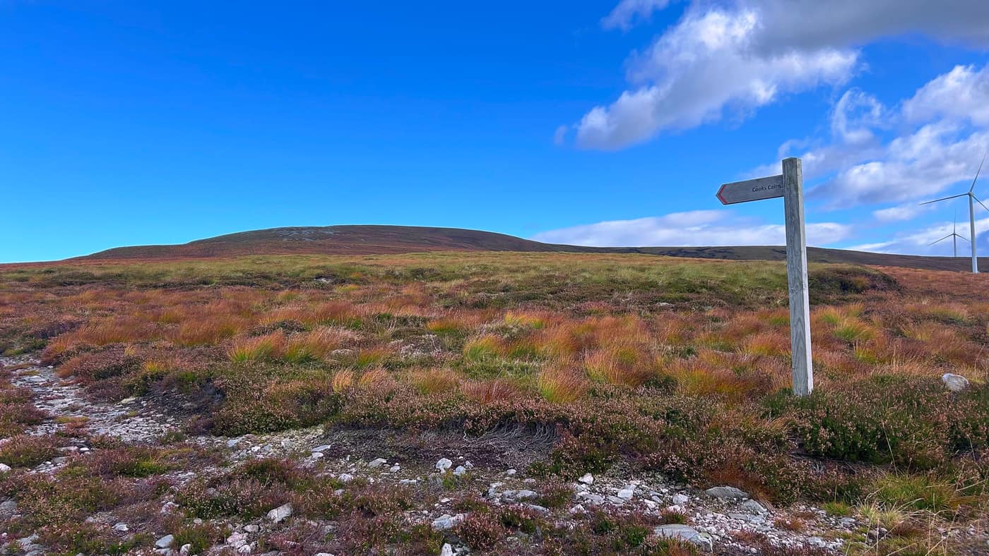The Cook's Cairn fingerpost with the hill's broad top ahead and a windmill at its back