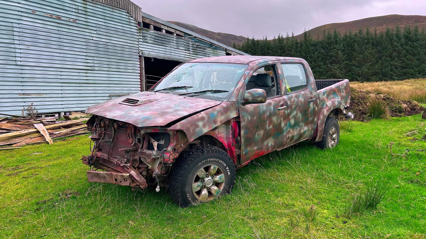 An abandoned camouflage-painted pickup beside the big barn