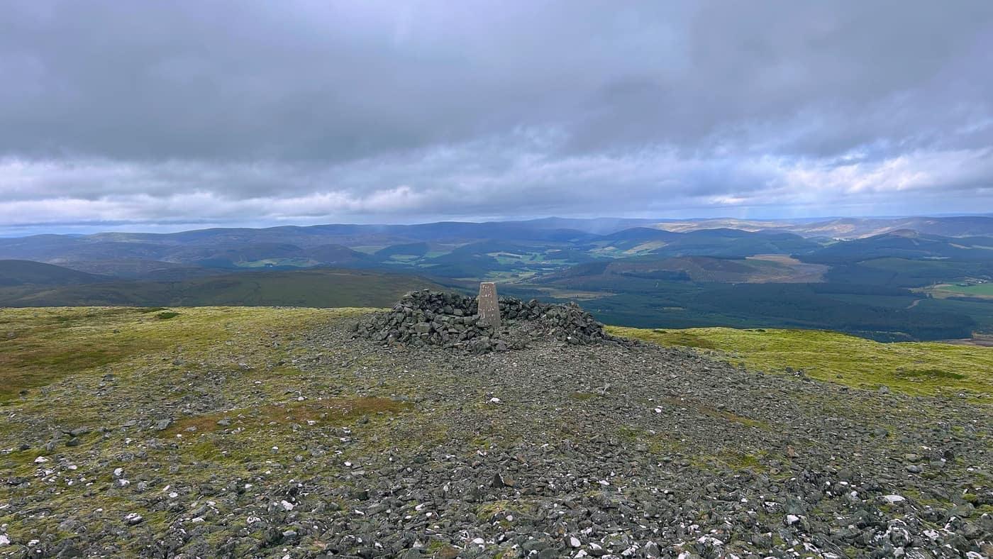 The summit plateau, the trig point on its cairn and the hills running away behind