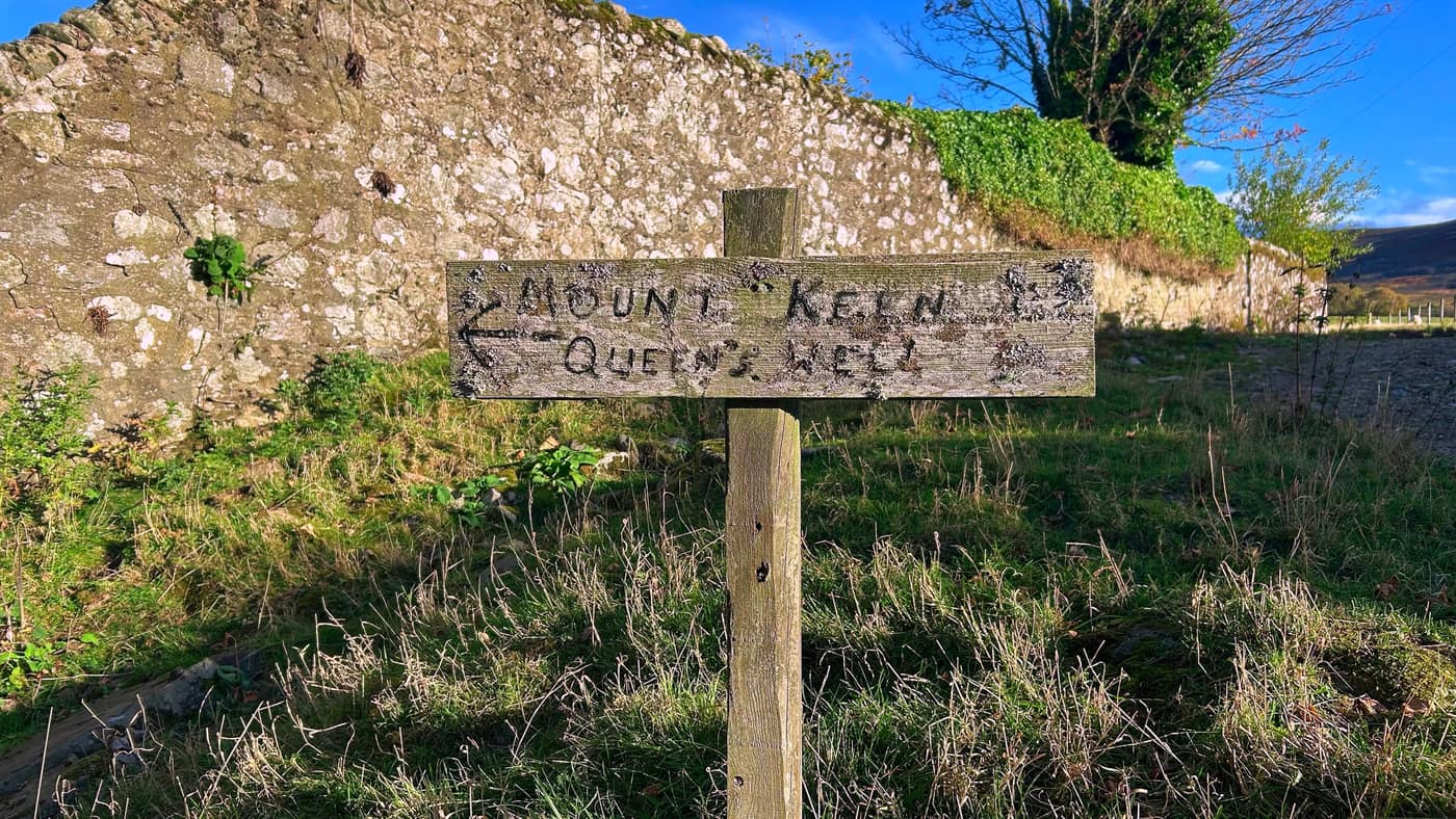 The old hand-painted wooden sign by the stone wall, pointing to Mount Keen and Queen's Well